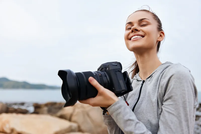 Smiling young female photographer holding a camera by the sea, dressed in a gray hoodie, conveying joy and creativity