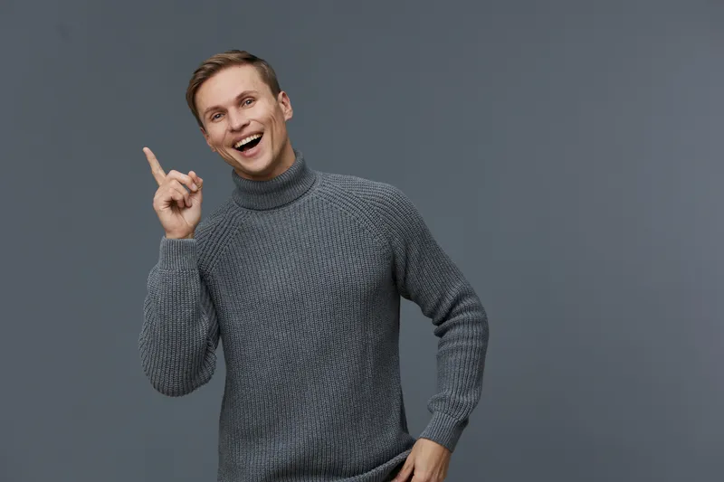 Smiling man in gray turtleneck sweater pointing up isolated on gray background happy mental health and gadgets concept