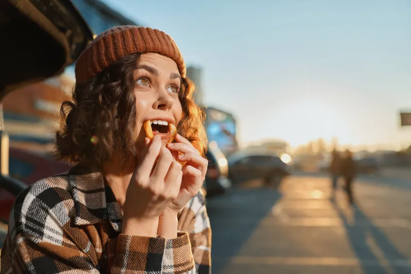 Woman eating a snack by a truck at sunset, wearing a beanie and curly hair, candid outdoors portrait capturing authenticity, candid lifestyle, golden hour glow and emotional storytelling.