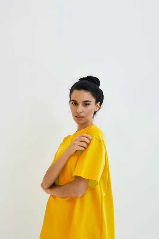 young woman wearing bright yellow oversized t-shirt standing with arms crossed against plain white wall, natural light, casual style confident expression