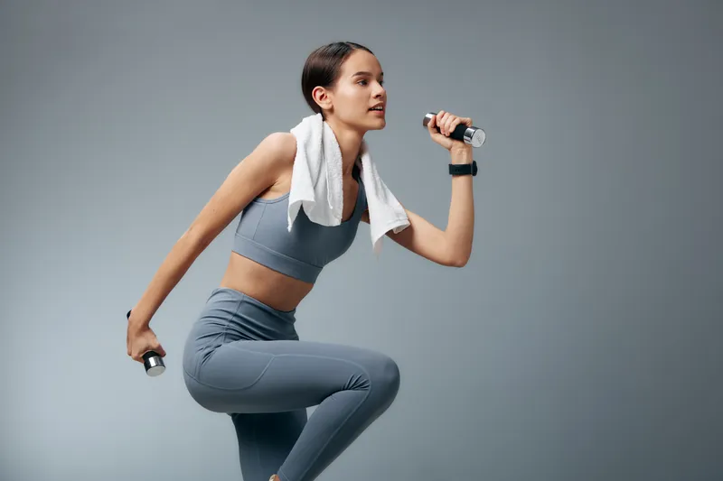 woman fitness sport workout training with dumbbells, towel draped over shoulder, focused athlete performing lunge for strength and cardio in a studio setting