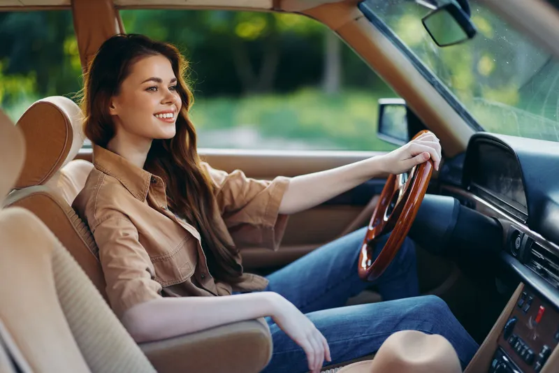 Young woman smiling in a stylish car interior, wearing a casual outfit with a warm color palette, evoking a sense of adventure and freedom in a natural setting.