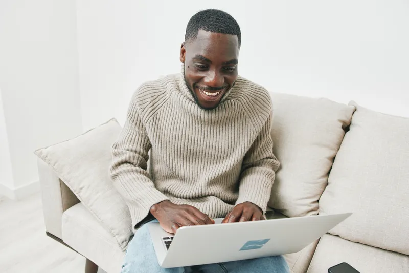 Happy Black man using a laptop in a cozy living room, wearing a warm sweater, showcasing a joyful expression and engaged in work or study