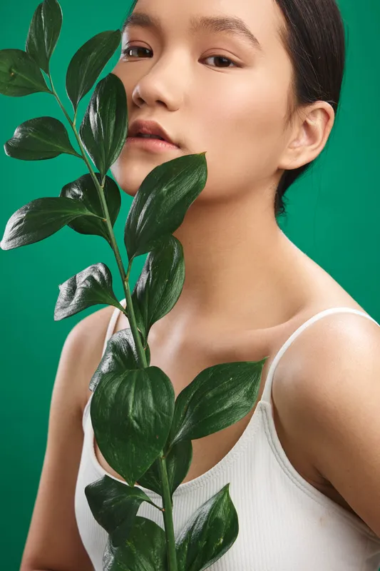 Asian woman with a calm expression holds a leafy stem against a vivid green backdrop, creating a fresh botanical portrait that emphasizes natural beauty, simplicity, and mindful styling.