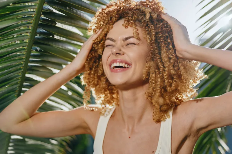 Smiling woman with curly hair enjoying tropical sunlight, surrounded by green palm leaves, wearing a light tank top, expressing happiness and relaxation outdoors.