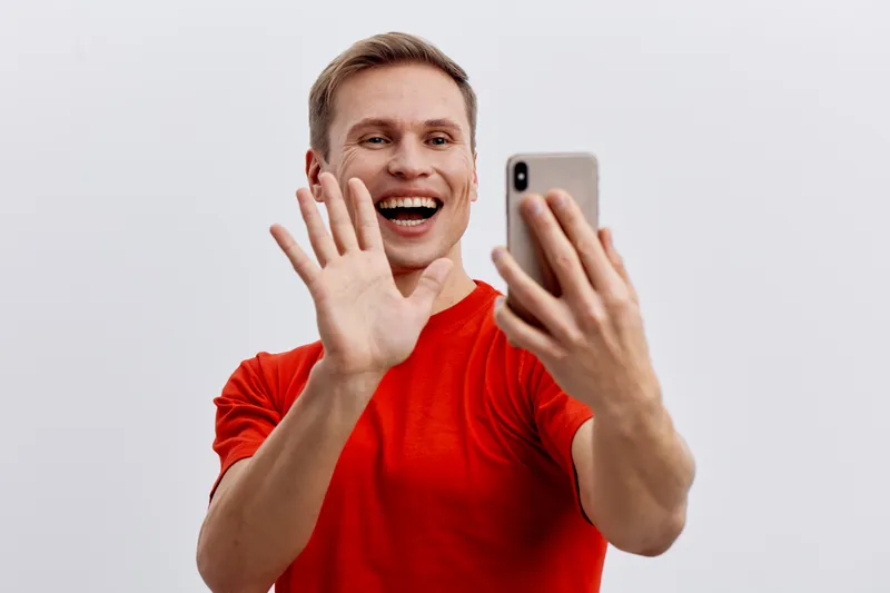Smiling young Caucasian man in red t-shirt taking selfie with smartphone isolated on light gray background, cheerful expression, casual style, lifestyle concept, happiness, communication, technology