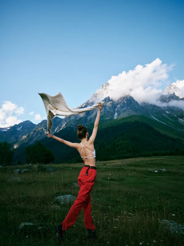 Dancer leaping in red pants with scarf in an open meadow beneath towering mountains, wind catching fabric, showcasing movement, freedom, and outdoor adventure