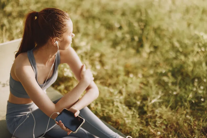 Young athletic woman listening to music in nature, enjoying a moment of relaxation and wellness during a sunny day, wearing sportswear and reflecting on life
