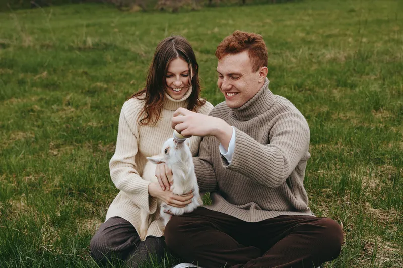 Happy couple outdoors with a goat kid in a green field, sharing smiles and gentle interaction, cozy sweaters, relaxed lifestyle, candid moment of companionship and nature.