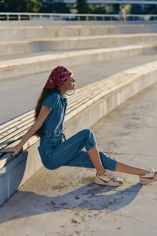 Lifestyle woman wearing modern boho-western denim street style outfit with red bandana and sneakers, sitting outdoors in warm sunlight, film color aesthetic.