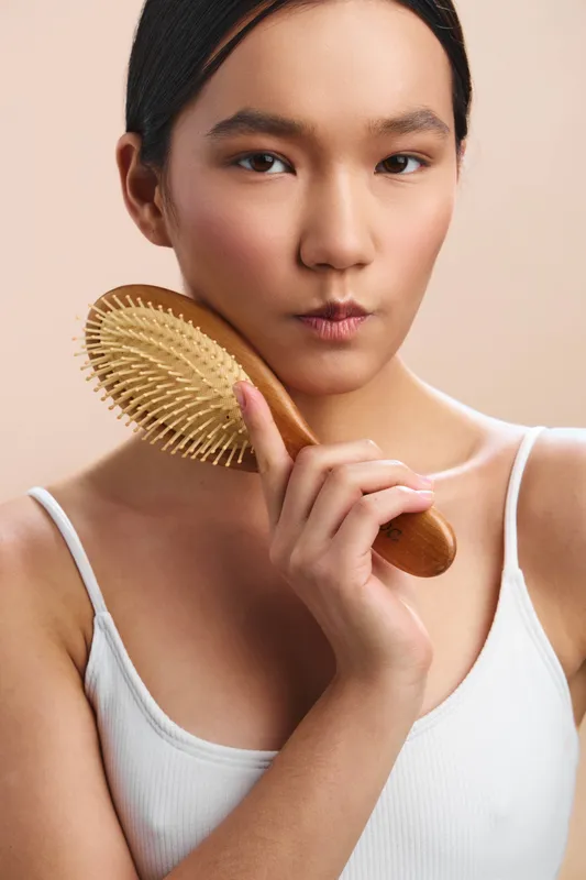 asian woman holds a wooden hairbrush in a soft studio portrait, calm and focused, with gentle lighting and a clean background, depicting a serene personal care moment.