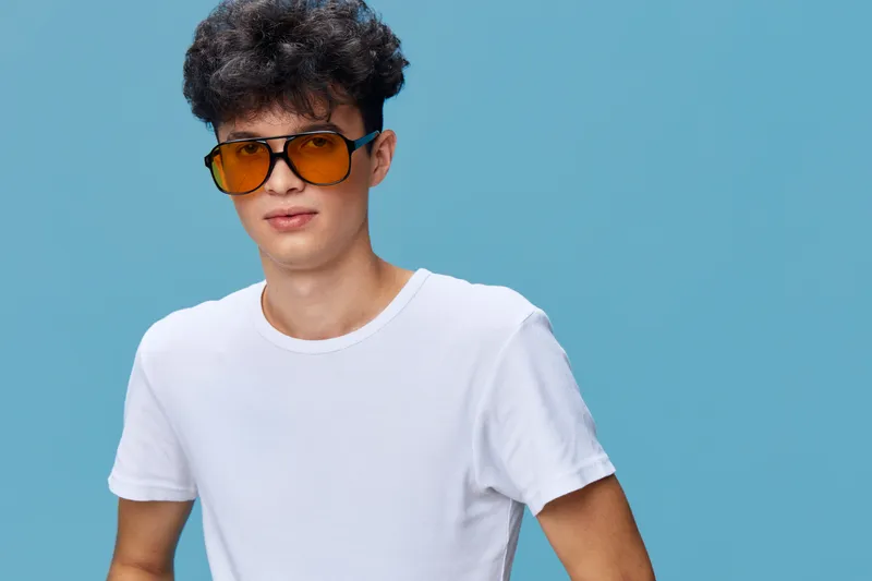 young man wearing yellow sunglasses and white t-shirt with curly hair posing confidently against plain blue background in casual style