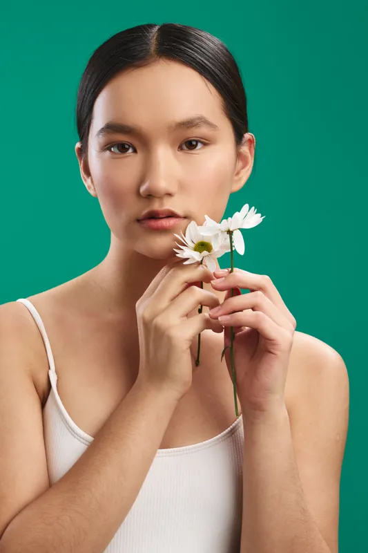asian woman softly holds a white flower near her face, standing against a vibrant green backdrop. she gazes at the camera with calm confidence and gentle charm.