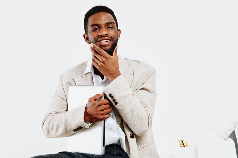 Smiling man with dark skin holds a tablet close to his chest, wearing a light beige sweater and white shirt, isolated on plain white background. People lifestyle concept, professional, confident, chee