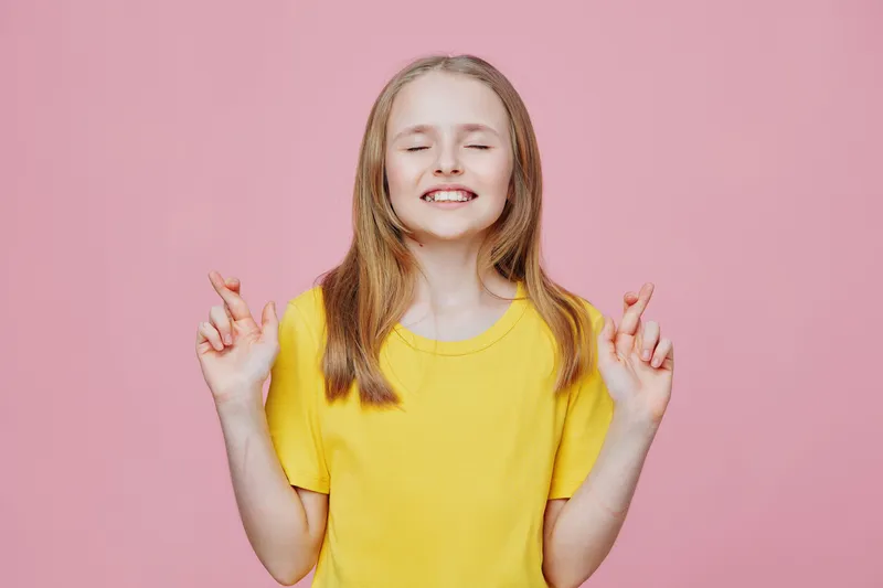 Cheerful girl with crossed fingers, wearing a bright yellow shirt, expresses hope and excitement against a pink background, ideal for themes of positivity and childhood