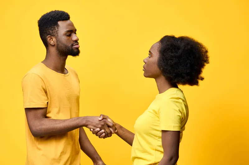Two young individuals, a man and a woman, shake hands against a vibrant yellow background, showcasing teamwork and agreement