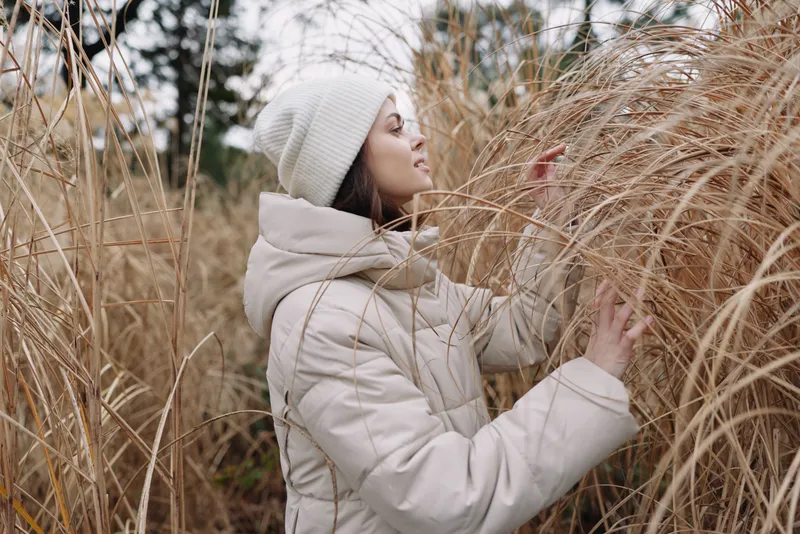 Woman in a warm coat and knit beanie stands among tall grass on a winter day, embracing outdoor nature and a calm, contemplative mood in a soft, muted scene
