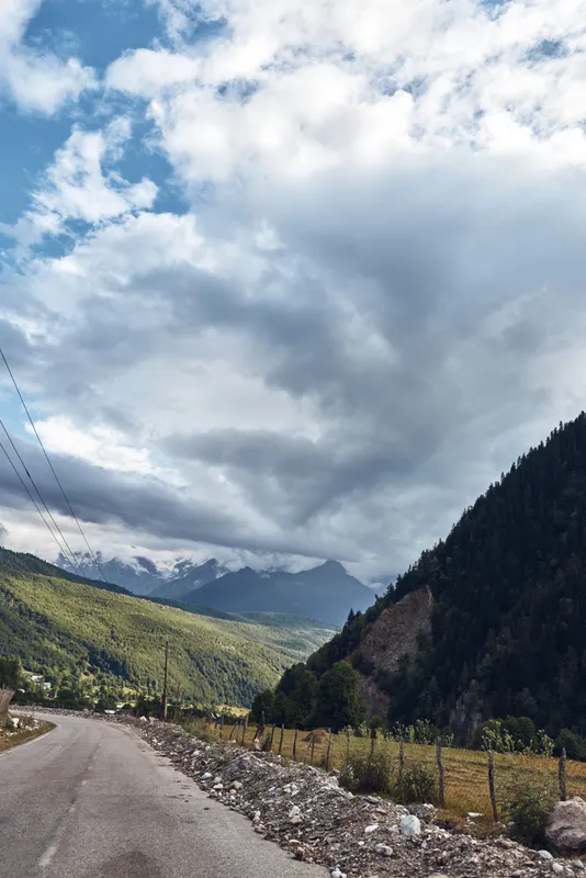 A winding road through mountains and landscape underneath a vast sky with clouds, fence lines along gravel shoulders within a valley, creating a rural countryside travel scene