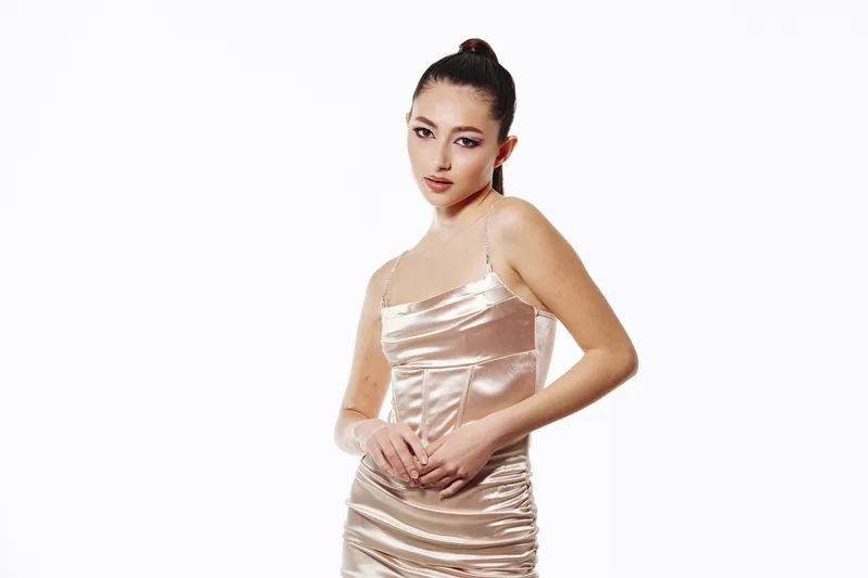 Young woman in a shiny satin dress poses confidently with sleek hair pulled back, wearing creative makeup. Studio shot on white background capturing fashion beauty and elegance.