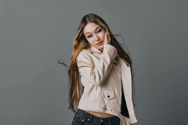 Young woman with long hair posing in light beige jacket and jeans on gray background. Fashion model expressing calm and relaxed attitude in studio shot with natural style.