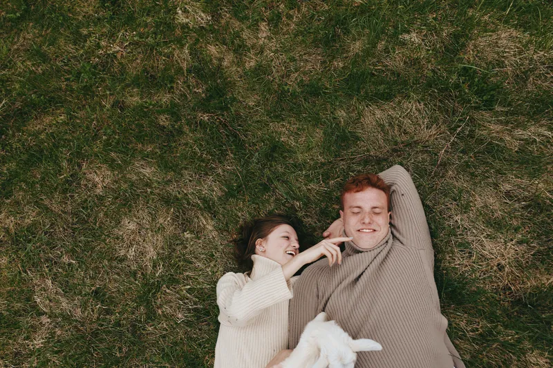 Young couple lying on grass wearing cozy sweaters enjoy playful moment with a white baby goat in a natural lifestyle setting outdoors in autumn.