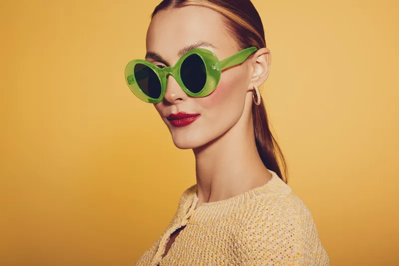 Stylish woman with green round sunglasses and red lipstick poses on yellow background. Fashion portrait with retro vibe, bright colors, and confident expression. Concept of summer style.