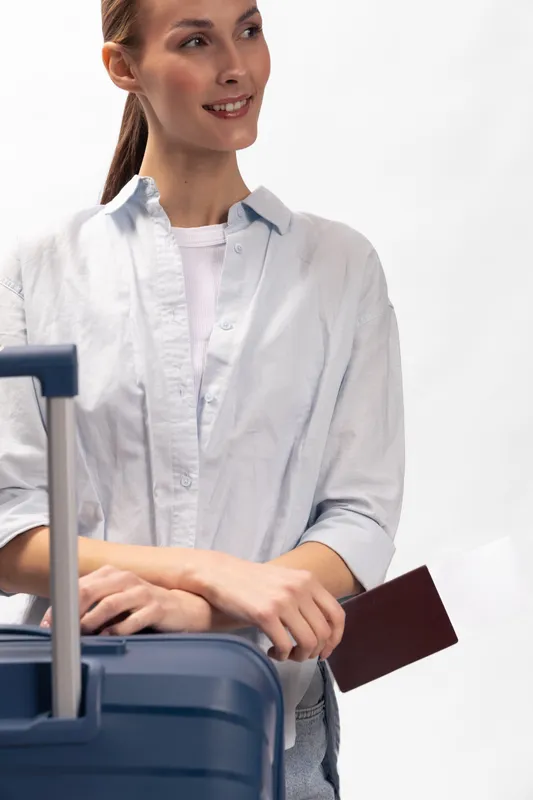Young woman holding passport stands near blue suitcase in casual white shirt smiling looking aside. Concept of travel preparation, vacation, tourism, and leisure lifestyle.