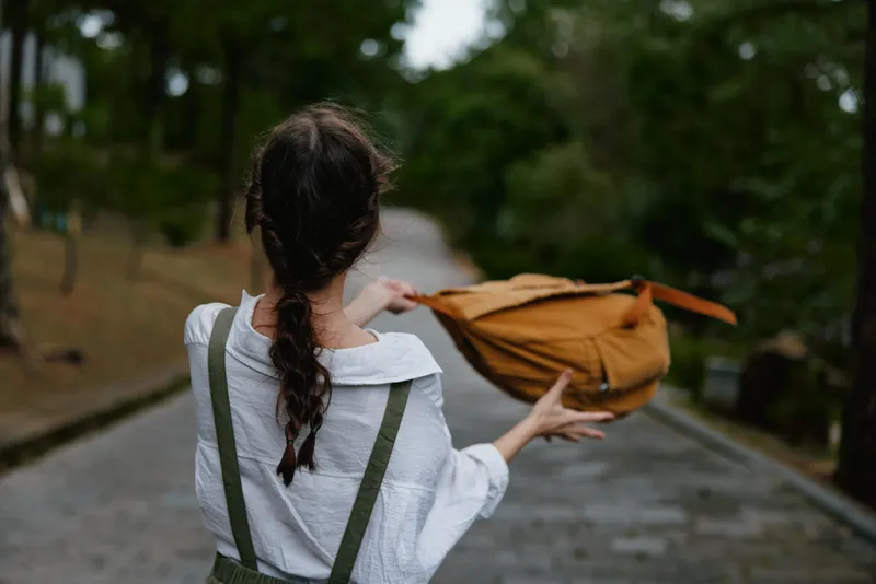 Young woman with long brown hair in a white shirt, joyfully tossing an orange backpack on a forest path, reflecting freedom and adventure in nature