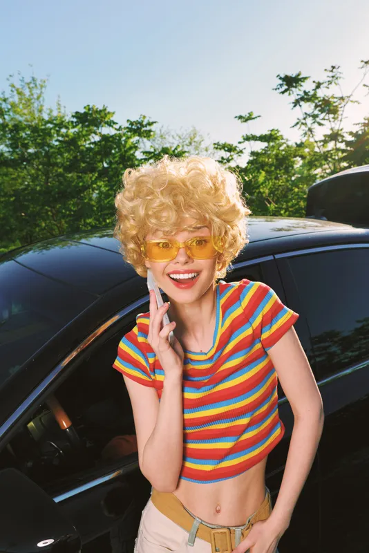 Young woman with curly blonde hair stands beside a dark car, wearing a colorful striped crop top and yellow sunglasses, smiling as she talks on her phone in a bright retro setting.
