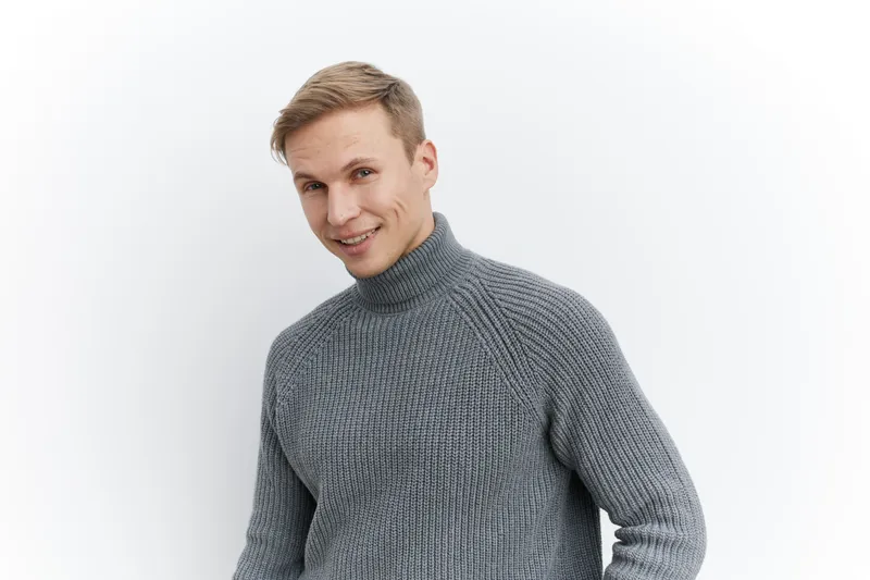Male smiling young adult with light brown hair wearing cozy gray knitted turtleneck sweater, looking at camera, cheerful facial expression, casual indoor studio setting, white background, fashion and 