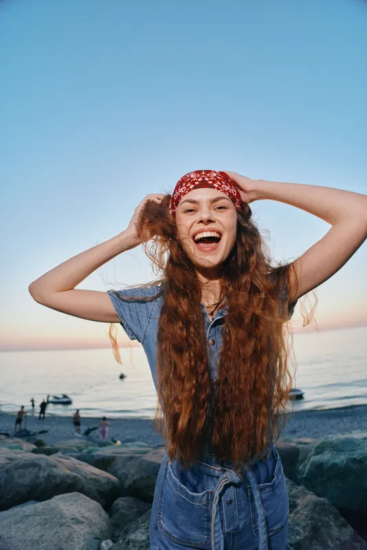 Lifestyle woman in modern boho-western denim street style laughing with long curly hair and red bandana by the beach at sunset in film color tones.