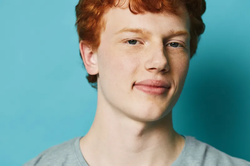 Young man with red hair and pale skin, smiling slightly with closed lips against blue background. Portrait of confident male wearing gray shirt, looking at camera with calm expression.