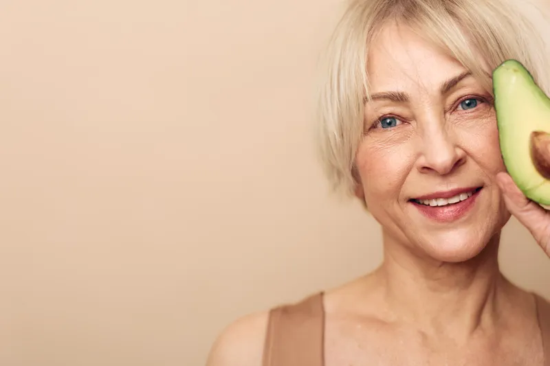 Smiling mature woman with short blonde hair holds fresh avocado half near face, natural skincare and healthy lifestyle concept with beige background and soft natural lighting.