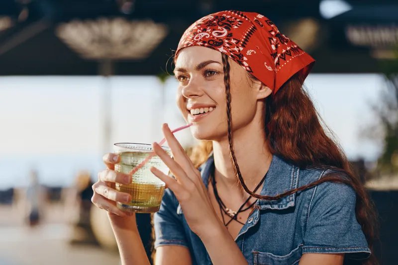 smiling woman in red bandana enjoying cold drink outdoors with casual denim top, warm sunlight, and relaxed summer vibe