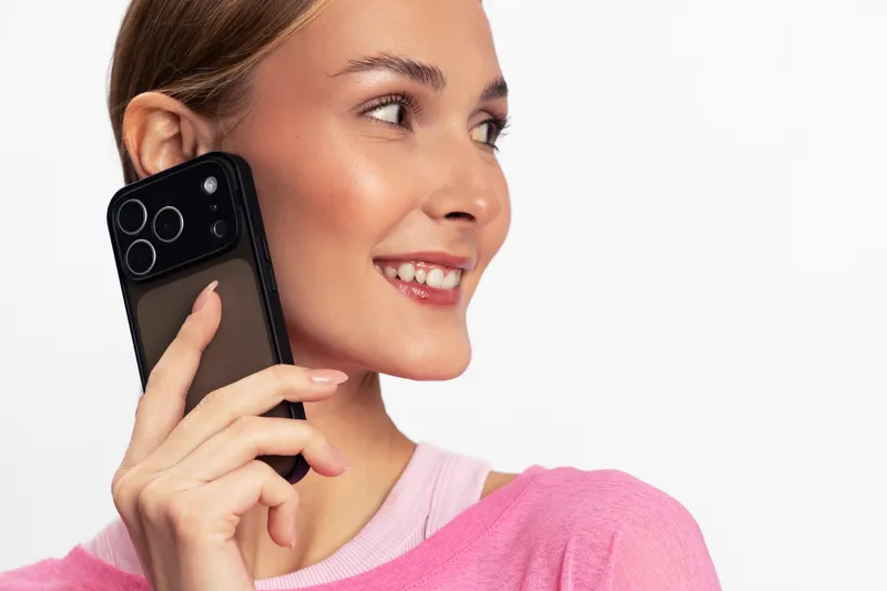 Young smiling woman holding smartphone near ear looking away on white background. Concept of communication, technology, positive emotions, and casual lifestyle with modern device.