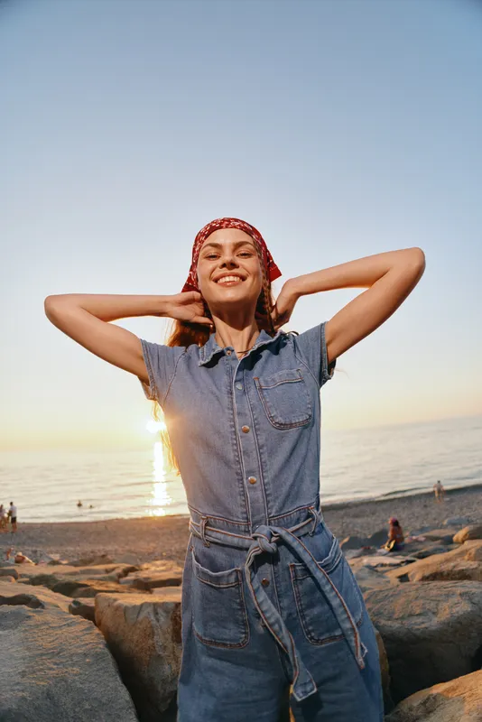 smiling woman with red bandana enjoys sunset on beach wearing denim jumpsuit with relaxed and joyful mood in warm natural light