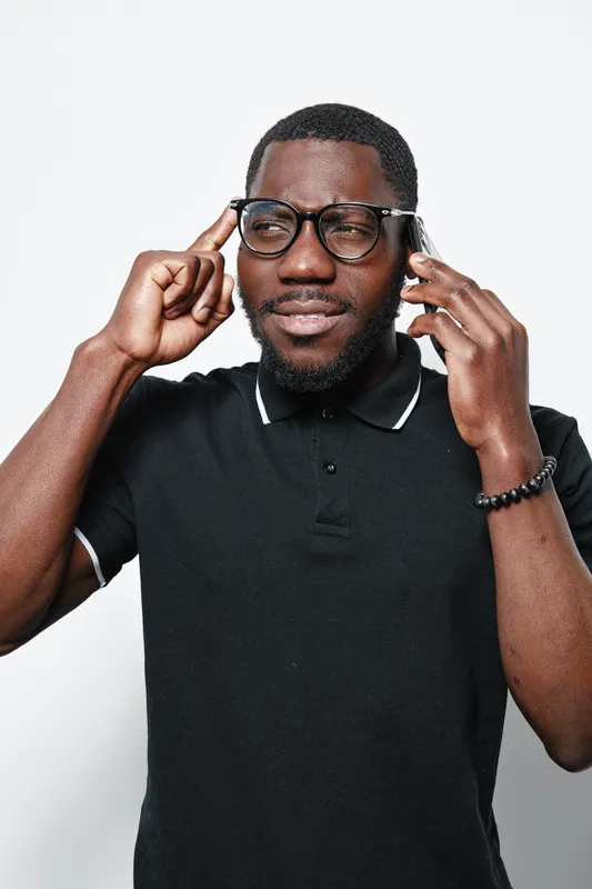 African American man with dark skin wearing glasses and black polo shirt thinking and talking on smartphone with focused expression on white background in studio portrait.