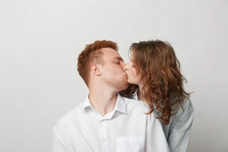 Young couple in white shirts kissing tenderly on a light background. Intimate moment of love and affection between man and woman with casual hairstyles and natural expressions.