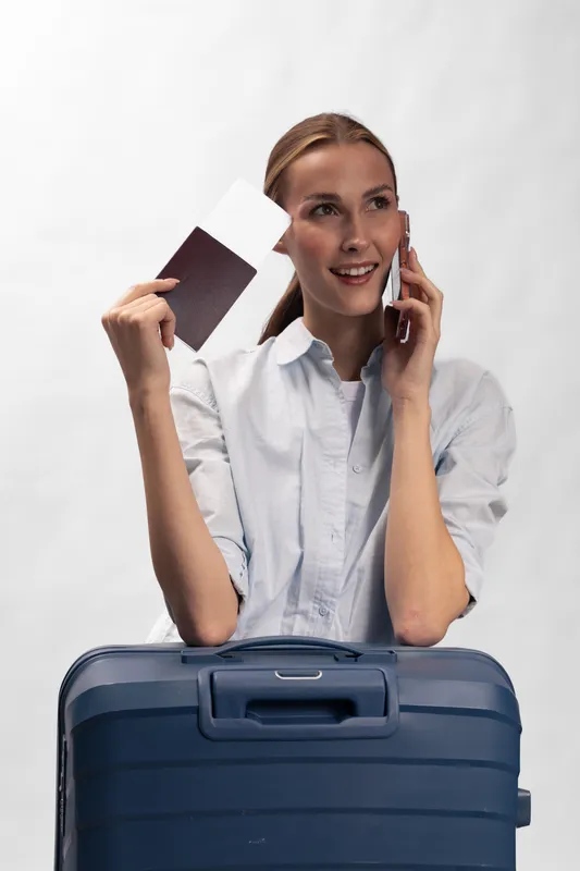 Young woman with a blue suitcase holding passport and tickets while talking on phone. Travel concept with happy female traveler preparing for a trip on white background.