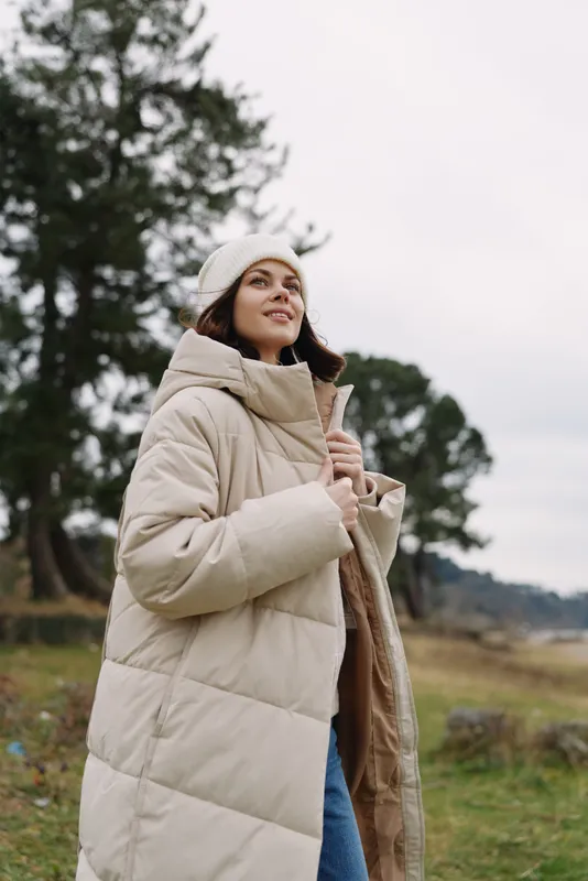 Woman wearing a beige puffer coat and white beret stands outdoors in a grassy field, looking up with a soft smile as wind blows through the chilly day.