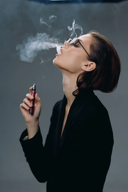 Young woman vaping with smoke exhaling, wearing glasses and black outfit, indoor lifestyle and modern concept.