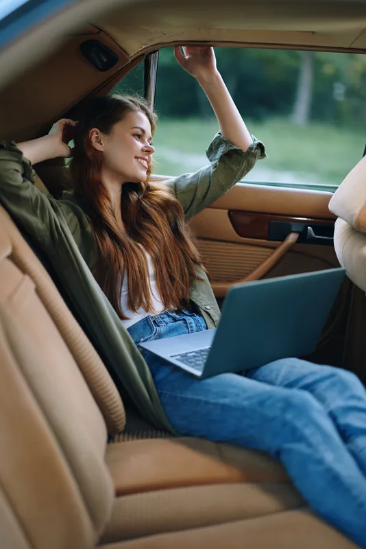 A young woman relaxes in a car, working on her laptop while smiling, expressing creativity and comfort, with a natural outdoor background and neutral color palette.