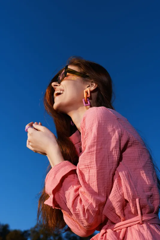 Joyful young woman in pink outfit smiling against a blue sky background, embodying happiness and fashion with vibrant colors in a sunny outdoor setting