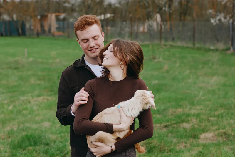 Affectionate couple outdoors in a park cuddle a small light dog, enjoying a relaxed lifestyle moment, close interaction and smiles, natural light, casual clothing, green grass backdrop, warm