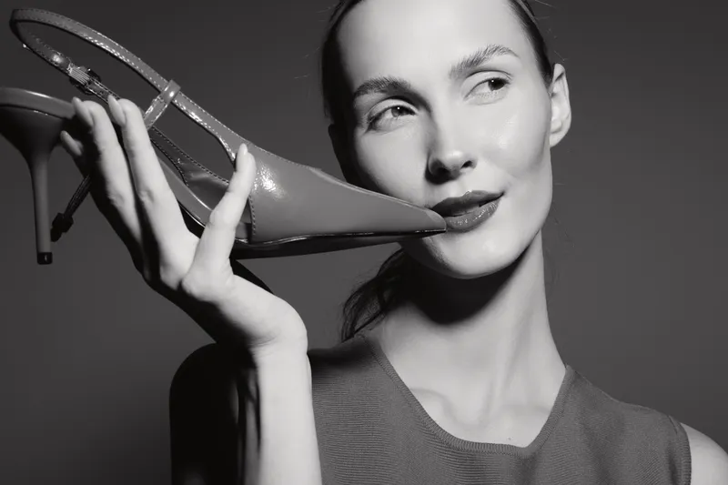 Slim white woman with pale skin in sleeveless top holds high heel shoe near face, looking sideways in black and white studio photo. Concept of fashion, elegance, and minimalism.