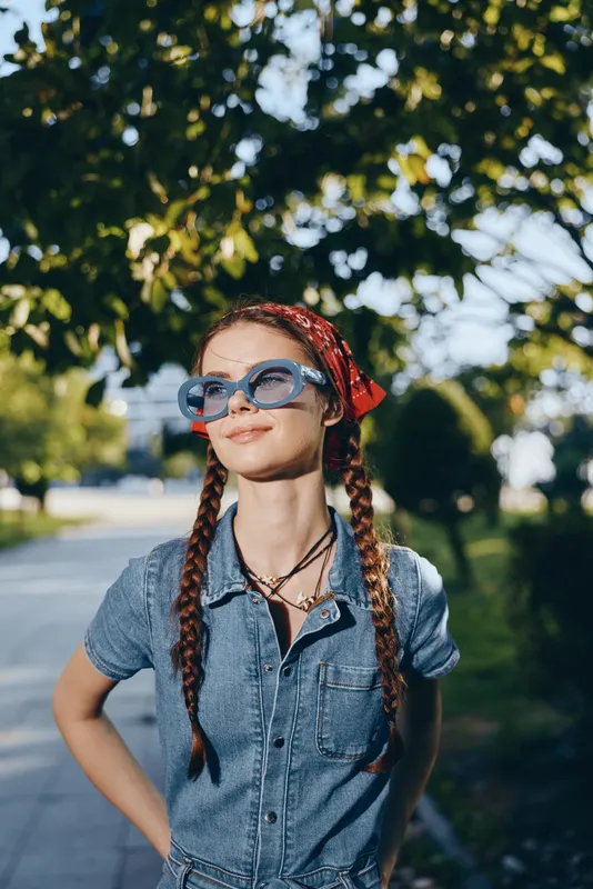smiling woman with braids wearing sunglasses and denim shirt enjoys sunny day outdoors in lush green park with relaxed and confident expression