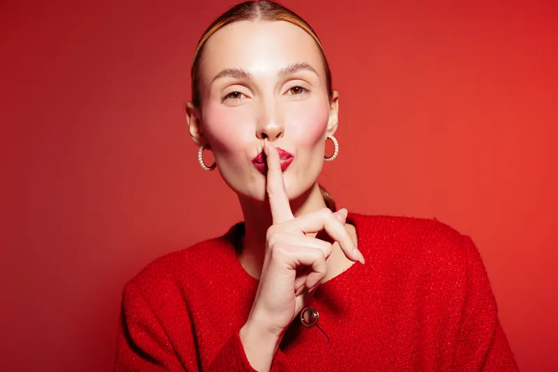 Woman in red sweater with finger on lips makes shushing gesture on red background. Concept of silence, secrecy, quiet, and calm expression in studio portrait photography.