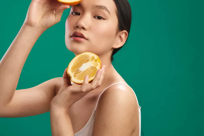 asian woman holds a freshly sliced orange near her face, a vibrant green backdrop emphasizes citrus freshness, bright lighting highlights smooth skin and moist lips, creating a healthy, refreshing
