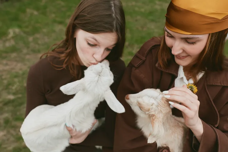 Two young women enjoy a sunny day outdoors while holding and interacting with white and beige baby goats in a casual lifestyle setting with green grass background.