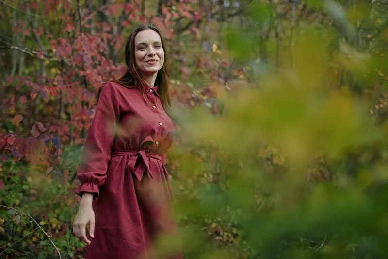 An authentic portrait of a thoughtful woman in a deep red dress standing among autumn foliage, conveying credibility through natural light and an unposed, sincere expression.